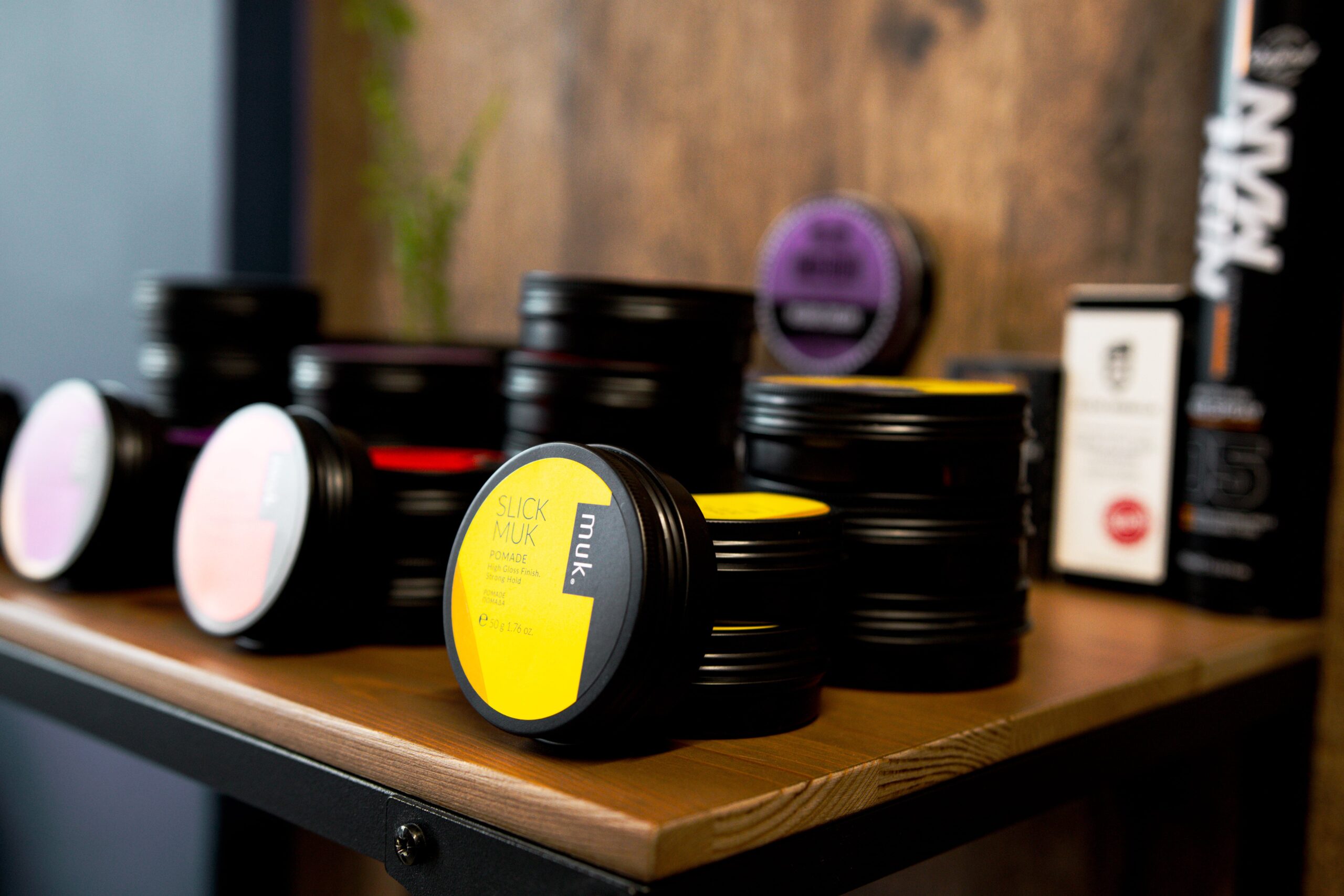 A collection of professional barber styling products, including Muk hair pomades, displayed on a wooden shelf in a barbershop in Larnaca.